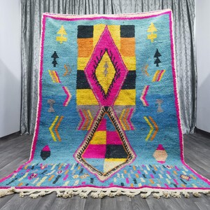 May include: A colorful Moroccan rug with a geometric pattern in shades of pink, yellow, blue, and black. The rug has a fringe border and is made of wool.