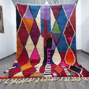 May include: A vibrant, hand-woven rug with a geometric diamond pattern. The rug features a rich palette of colors including red, blue, orange, purple, and white. It has a fringed edge and is likely made of wool or a similar material.