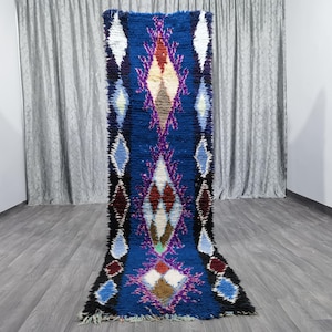 May include: A long, narrow, blue and purple Moroccan rug with a diamond pattern. The rug is made of wool and has a hand-woven texture.