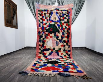 May include: A colorful Moroccan rug with a geometric pattern in shades of pink, blue, green, and red. The rug has a fringe border and is made of wool.