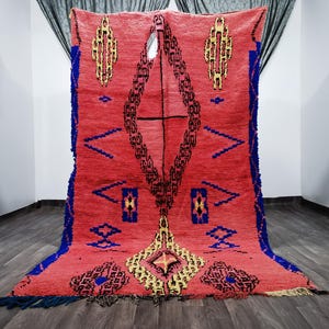 May include: A red Moroccan rug with a geometric pattern in shades of blue, yellow, and black. The rug has a fringe border and is woven with a high pile.