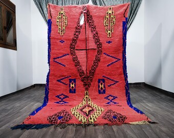 May include: A red Moroccan rug with a geometric pattern in shades of blue, yellow, and black. The rug has a fringe border and is woven with a high pile.