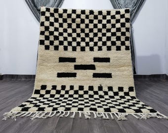 May include: A cream-coloured rug featuring a black chequered pattern at the top and bottom, with black rectangular shapes in the centre. The rug has a fringe along the bottom edge. This geometric design rug is suitable for home decor.
