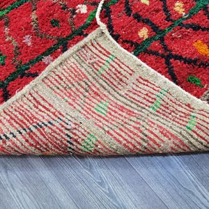 May include: A close-up of a red rug with a geometric pattern in green and black. The underside of the rug is visible, showing a striped pattern in red, white, and green. The rug is on a gray wooden surface.