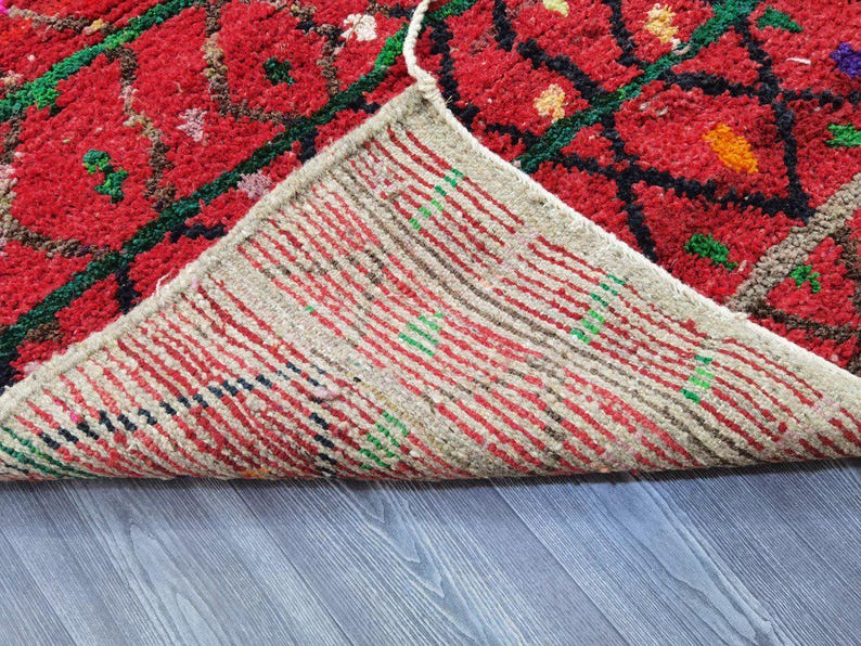 May include: A close-up of a red rug with a geometric pattern in green and black. The underside of the rug is visible, showing a striped pattern in red, white, and green. The rug is on a gray wooden surface.