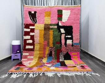 May include: A colorful Moroccan rug with geometric patterns in shades of pink, red, yellow, green, and black. The rug has a fringe border and is made of wool.