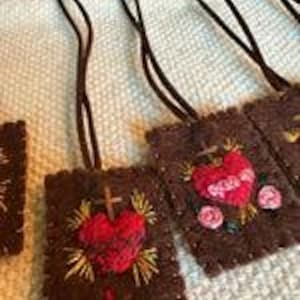 May include: Five brown felt pendants with embroidered images of a heart with thorns and a cross. The hearts are red and the crosses are gold. The pendants are strung on black cords.