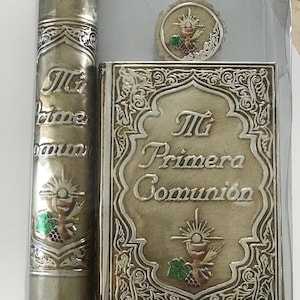 May include: A silver-toned First Communion set, including a book, candle, and a small round emblem. The book and candle feature the text "Mi Primera Comunión" and decorative religious symbols. The set is presented in a clear plastic case.