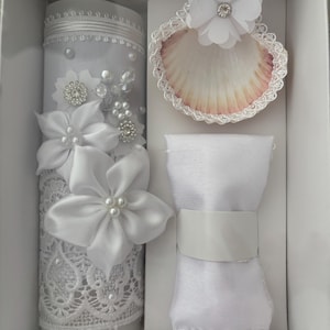 Könnte beinhalten: Ein weißes Taufset in einer Box. Das Set enthält eine dekorierte Kerze mit Spitze und floralen Akzenten, eine Muschel mit einer Blume und ein weißes Tuch. Die Kerze ist mit weißen Satinblüten, Perlen und Strass verziert.