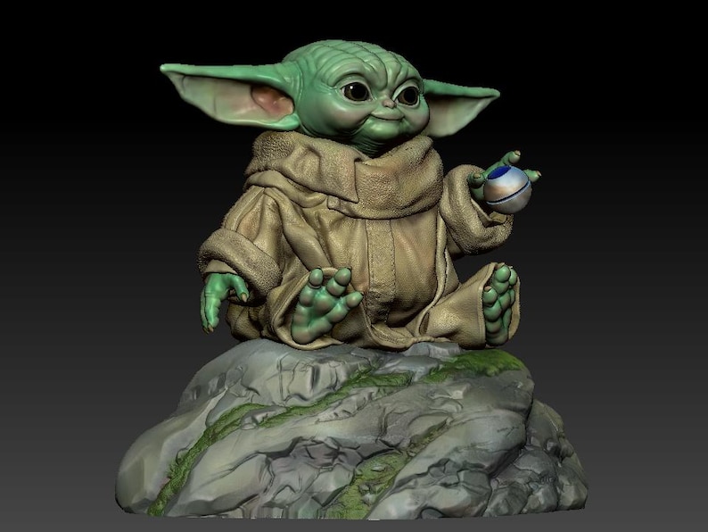 May include: A green and brown figurine of a character from the Star Wars franchise, sitting on a rock with a small sphere in its hand.
