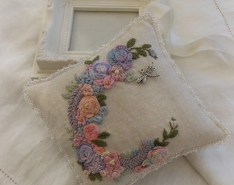 May include: A small, cream-coloured pillow with a beaded trim and a delicate embroidered floral design in shades of pink, purple, and green. A silver dragonfly charm is attached to the pillow.