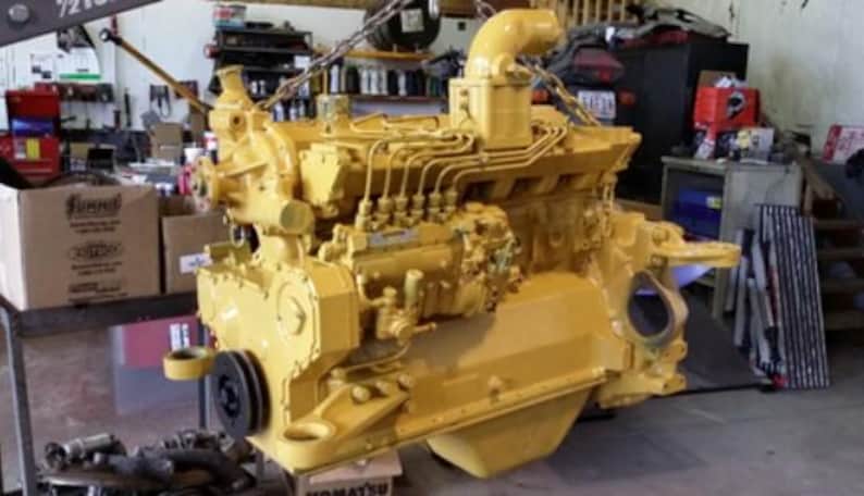 May include: A large, yellow Caterpillar diesel engine suspended by a chain hoist. The engine has numerous pipes, tubes, and a black pulley. The background shows a cluttered workshop with boxes and tools.