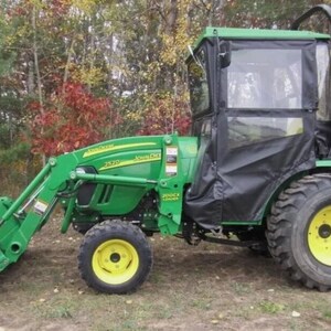 Op de afbeelding: Een groene John Deere tractor met een voorlader en een zwarte cabine. De tractor heeft gele wielen en staat geparkeerd op een grasveld met bomen op de achtergrond.