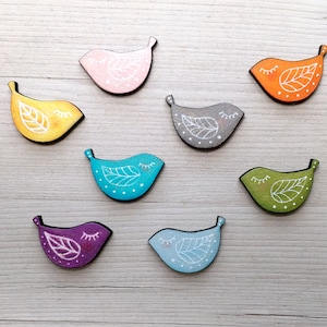 Può includere: Otto ornamenti di uccelli in legno dipinti in vari colori, tra cui giallo, arancione, rosa, blu, verde, viola e grigio. Ogni uccello ha un disegno di foglia bianca e gli occhi chiusi.
