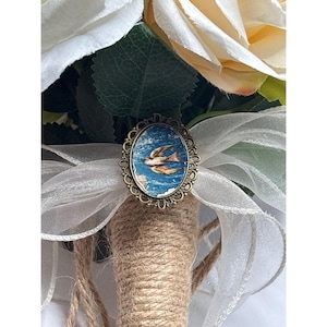 May include: A bridal bouquet featuring cream-colored roses and white ribbon. The bouquet is accented with a vintage-style oval brooch depicting a bird in flight against a blue sky. The stems are wrapped in twine.