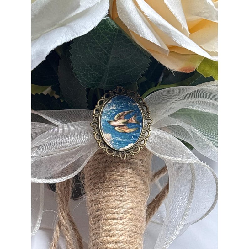 May include: A bridal bouquet featuring cream-colored roses and white ribbon. The bouquet is accented with a vintage-style oval brooch depicting a bird in flight against a blue sky. The stems are wrapped in twine.