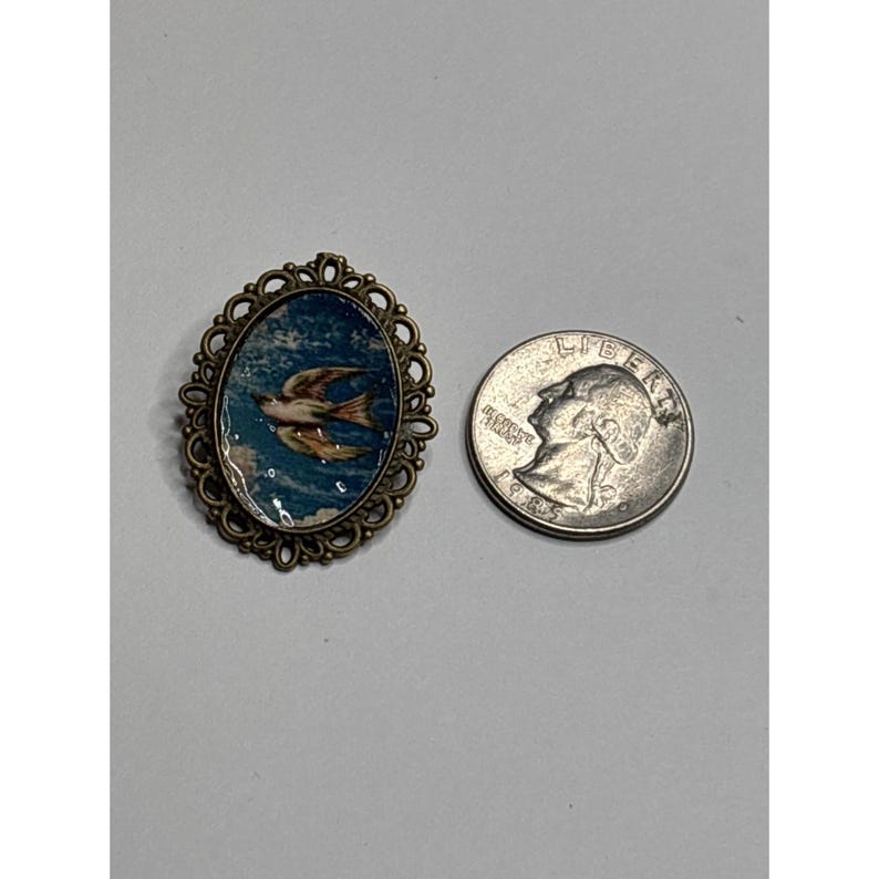 May include: An oval-shaped vintage brooch with a bird design. The brooch features a detailed, ornate bronze-colored frame surrounding a painted image of a bird in flight against a blue sky. A silver quarter is next to the brooch for size comparison.