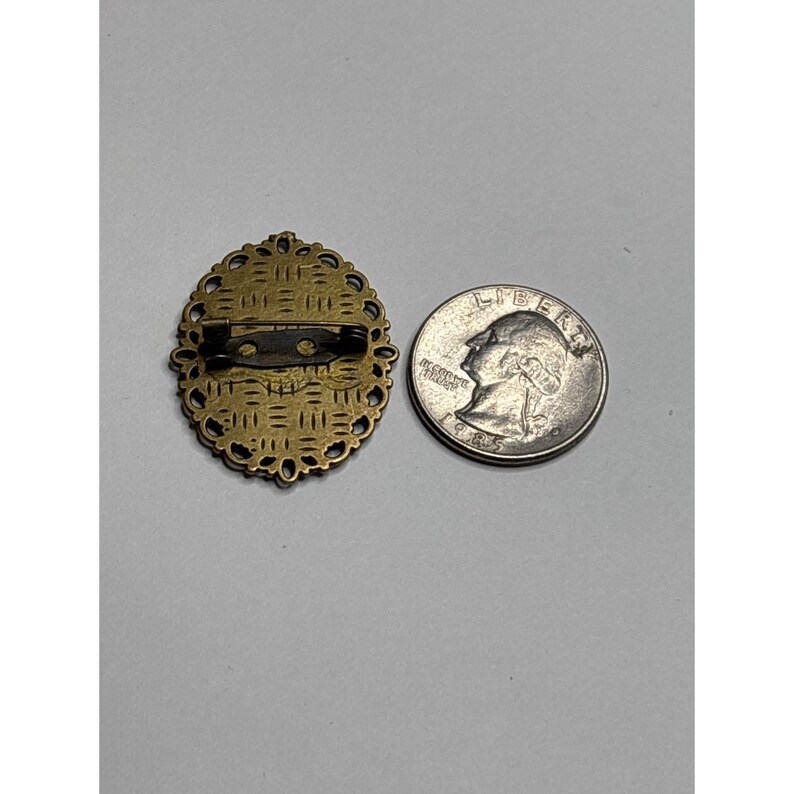 May include: An antique brass-toned oval brooch with an ornate filigree design. The brooch has a pin closure on the back. A US quarter is next to the brooch for size comparison.