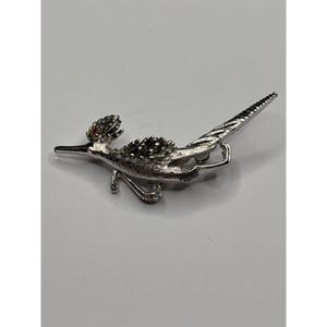 May include: A silver-toned roadrunner brooch with a detailed design. The bird has a long beak, a spiky crest, and a long tail. The brooch is likely made of metal and features a red accent on the head.