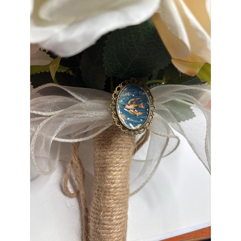 May include: A bridal bouquet featuring cream-colored roses and greenery. The stems are wrapped in twine and adorned with a vintage-style brooch. A sheer white ribbon adds a delicate touch to the arrangement. The brooch has a blue background with a bird.