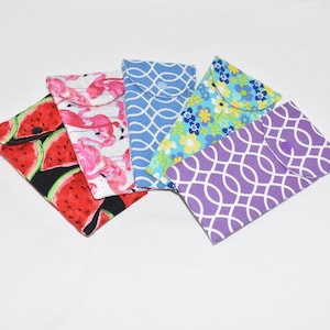 Könnte beinhalten: Fünf Stoffbeutel mit verschiedenen Mustern. Die Beutel sind aus Baumwolle und haben einen Knopfverschluss. Die Muster umfassen Wassermelone, Flamingos, blau-weißes geometrisches Muster, Blumenmuster und lila-weißes geometrisches Muster.