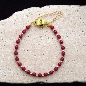 May include: A delicate bracelet featuring small, round, deep red beads interspersed with gold-colored beads. The bracelet has a gold-colored clasp and chain. The jewelry is displayed on a textured, light-colored surface.