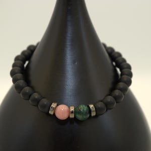 Puede incluir: Un brazalete de cuentas negras con tres cuentas de piedras preciosas: una cuenta rosa, una cuenta plateada y una cuenta verde.