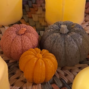 May include: Three small decorative pumpkins, one orange, one pink and one grey, are arranged on a woven basket surface.