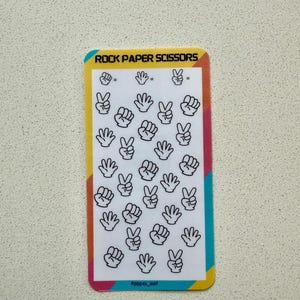 May include: A rectangular sticker with a white background featuring black line drawings of hands in rock, paper, and scissors gestures. The sticker has a colorful border and the text "ROCK PAPER SCISSORS" at the top.