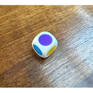 May include: A white, multi-colored die with purple, blue, and yellow circles on its faces. The die is set against a wooden background, with the word "STYLES" visible at the top of the image.