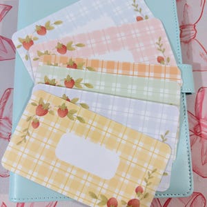 May include: A pale blue planner with a tab closure, holding several colourful cards. The cards feature a plaid pattern in pastel shades, decorated with strawberry and leaf illustrations. The cards have a blank white space for writing.