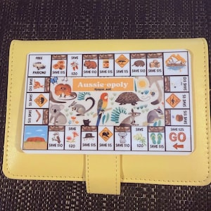 May include: A yellow leather wallet featuring a game board design. The game board depicts Australian animals and landmarks, with the text "Aussie-opoly". The wallet has a snap closure.