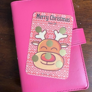 May include: A pink leather planner with a Christmas-themed sticker featuring a cartoon reindeer and the text "Merry Christmas" and "Saves: £30".
