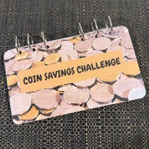 May include: A spiral-bound notebook with a coin-themed cover and the text "COIN SAVINGS CHALLENGE" on a tan banner. The cover features a collage of coins in various colours, including silver and gold. The notebook is open, revealing the coin design.