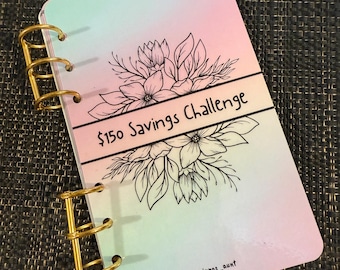 May include: A pastel-colored notebook with gold binder rings. The cover features a floral design and the text "$150 Savings Challenge". The notebook has a gradient of pink, blue, and green.