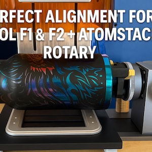 May include: A black rotary laser engraving machine with a cylindrical object featuring a colorful dragon design. The text "PERFECT ALIGNMENT FOR xTool F1 & F2 + ATOMSTACK R1 ROTARY" is displayed in white. The machine is on a black surface.
