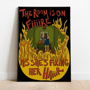 May include: A framed art print with a black border. The artwork shows a woman in a blue dress fixing her hair in a mirror, surrounded by flames. Text reads "THE ROOM IS ON FIIIIRE AS SHE'S FIXING HER HAIIIR".