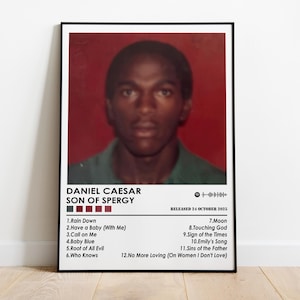 May include: A framed poster featuring a portrait of Daniel Caesar with the text "SON OF SPERGY." The poster includes a tracklist with song titles like "Rain Down" and "Moon," and the release date of October 24, 2015. The background is red.