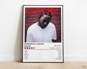 May include: A framed poster featuring Kendrick Lamar's album "DAMN." The poster has a photo of the artist in a white shirt against a brick wall. The album title is in red, with the tracklist and release date below.