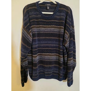 May include: A blue, brown, and tan striped sweater with a crew neck. The long-sleeved sweater is hanging on a white hanger against a white wall. The sweater has a textured knit pattern.
