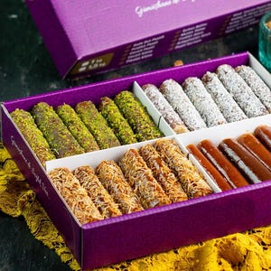 Op de afbeelding: Een paarse geschenkdoos gevuld met verschillende Turkse lekkernijen. De doos bevat vier verschillende soorten snoep, waaronder pistache-bedekte, geraspte tarwe en een karamel-achtig snoepje. De doos staat op een geel en bruin gepatroneerd oppervlak.