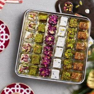 May include: A silver tray filled with assorted Turkish delights. The tray is divided into squares, each containing a different flavor of Turkish delight. The Turkish delights are a variety of colors, including white, pink, green, and brown. The tray is sitting on a white surface.