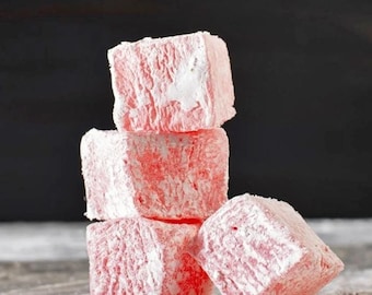 May include: A stack of five pink Turkish delight candies dusted with powdered sugar. The candies are square-shaped and have a soft, textured appearance. The background is a dark gray, providing contrast to the light pink sweets.