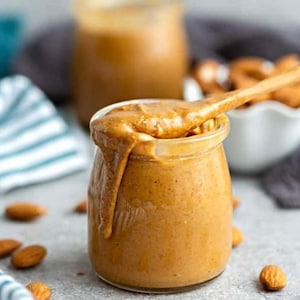 May include: A glass jar filled with smooth, creamy almond butter. The jar is on a gray surface with almonds scattered around it. A wooden spoon is partially submerged in the jar, with a dollop of almond butter on the spoon.