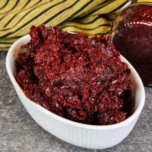 Op de afbeelding: Een witte ovale kom gevuld met een donkerrood, grof voedsel. Het voedsel heeft een versnipperde textuur en lijkt op een relish of spread. Een pot van hetzelfde voedsel is zichtbaar op de achtergrond, samen met een geel en zwart gestreepte doek.