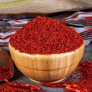 Può includere: Una ciotola di legno piena di fiocchi di peperoncino rosso macinato. La ciotola è su uno sfondo di tessuto a righe.