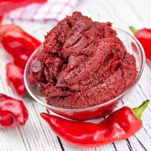 Op de afbeelding: Een glazen kom gevuld met dieprode tomatenpuree. Verschillende felrode chilipepers zijn rond de kom verspreid op een wit houten oppervlak. De pasta heeft een dikke, getextureerde uitstraling.