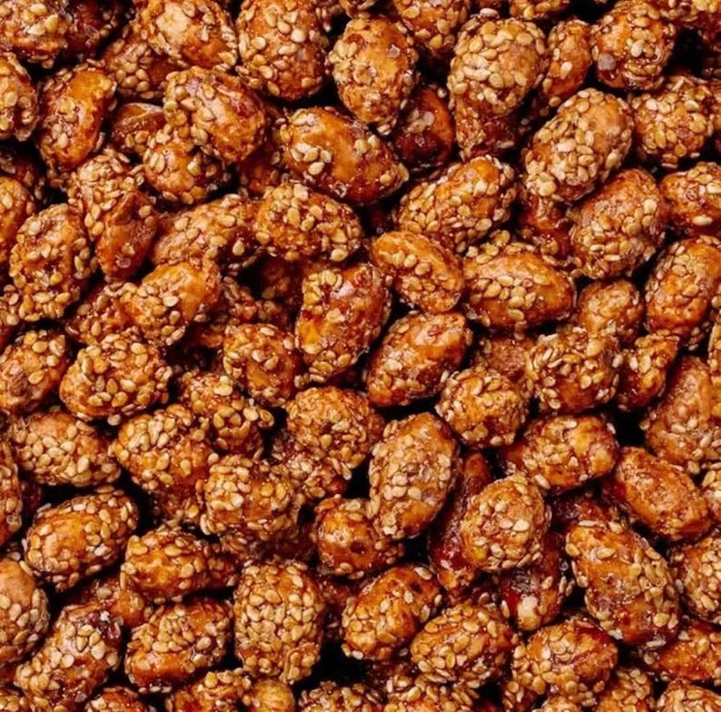 Op de afbeelding: Close-up van een grote stapel geroosterde pinda's bedekt met sesamzaadjes. De pinda's zijn lichtbruin van kleur en de sesamzaadjes zijn donkerbruin van kleur.
