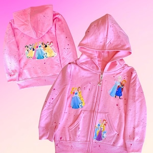 K&ouml;nnte beinhalten: Rosaer Kinder-Rei&szlig;verschluss-Hoodie mit Spr&uuml;hfarben-Design. Der Hoodie zeigt verschiedene Disney-Prinzessinnen auf der Vorder- und R&uuml;ckseite.