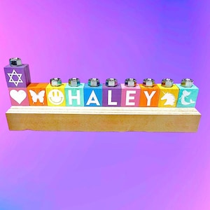 May include: A rainbow-coloured wooden menorah with the name "HALEY" spelled out on the front. Each block has a different design, including a Star of David, a heart, a butterfly, a smiley face, a unicorn, and a mermaid.